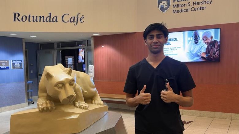 Faaiq Rizwan, wearing scrubs and giving two thumbs up, stands near a Nittany Lion statue at Penn State Health Milton St. Hershey Medical Center